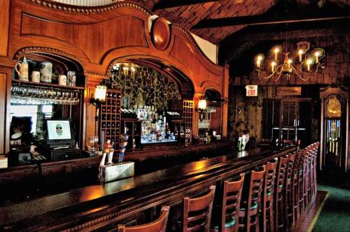 A wooden bar with a long counter, shelves of liquor, and a chandelier in a cozy, dimly lit pub interior.