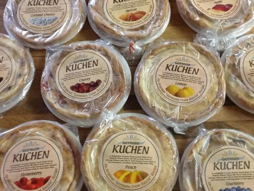 A variety of German kuchen pastries in circular containers, labeled with flavors like cherry, lemon, and peach.