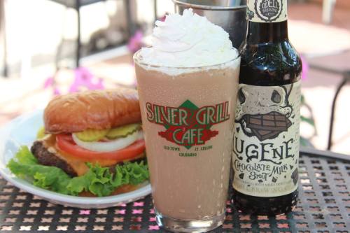 A glass of chocolate milk stout topped with whipped cream, alongside a burger with lettuce, tomato, and pickles.