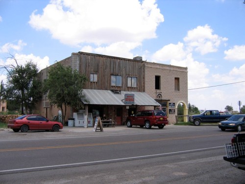 A rustic two-story building with a wooden facade, featuring a store front and parked cars on a rural road.