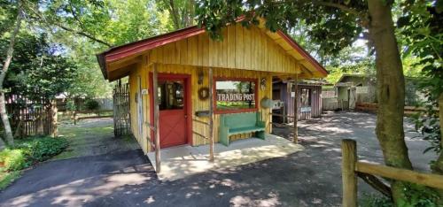 A wooden trading post with a red door and green bench, surrounded by trees and a gravel path.