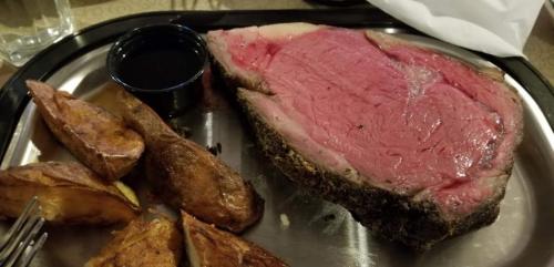 A plate with a large slice of rare prime rib and seasoned potato wedges, accompanied by a small cup of sauce.