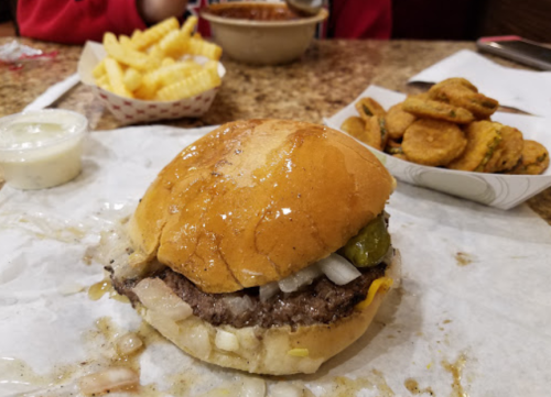 A close-up of a burger with onions and pickles, accompanied by fries and a bowl of chili in the background.