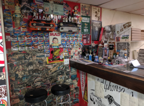 A vintage-themed bar area with a collage of car memorabilia, posters, and two stools in front of a wooden counter.