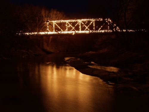 A bridge adorned with lights reflects in a calm river at night, surrounded by dark trees.