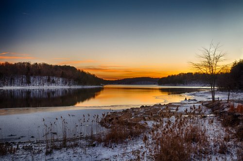 A serene winter landscape at sunset, featuring a calm lake, icy shores, and trees silhouetted against the colorful sky.
