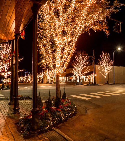 A street corner at night, adorned with glowing lights on trees and festive decorations, creating a warm atmosphere.
