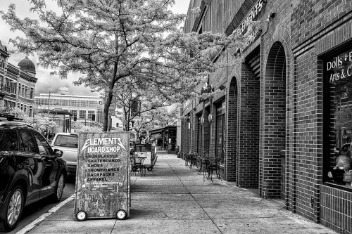 Black and white street scene featuring a board shop sign, trees, and outdoor seating along a brick building.
