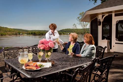 A server brings drinks to two women at a table with flowers and snacks, overlooking a scenic river view.