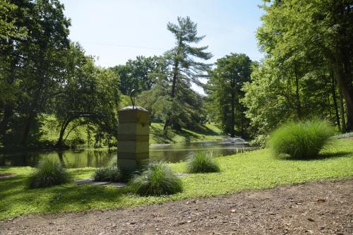 A serene park scene featuring a stone structure by a pond, surrounded by lush greenery and tall trees under a clear sky.