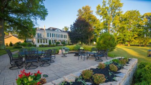 A spacious patio with seating, surrounded by lush greenery and a large house in the background under a clear blue sky.