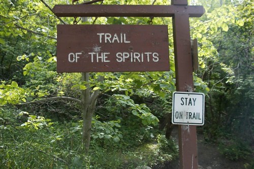 A wooden sign reading "Trail of the Spirits" next to a sign that says "Stay on Trail," surrounded by greenery.
