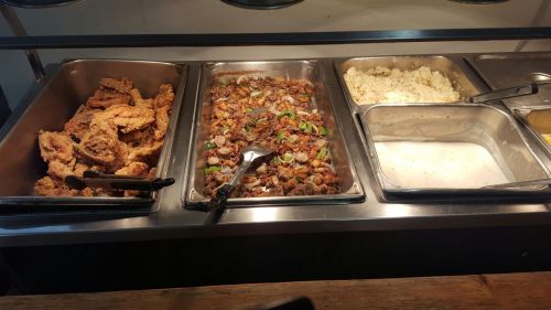 A buffet display featuring fried chicken, mixed meat with vegetables, mashed potatoes, and gravy in metal trays.