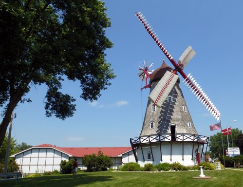 A large windmill with red and white blades stands beside a green lawn and a building, under a clear blue sky.