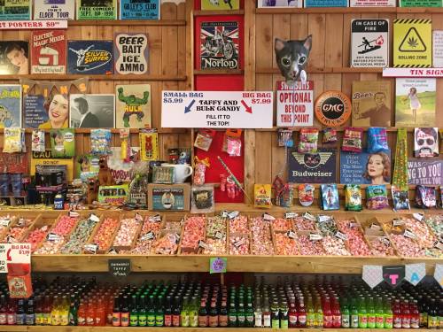 A colorful display of candy, snacks, and drinks in a rustic wooden shop, with signs for pricing and products.