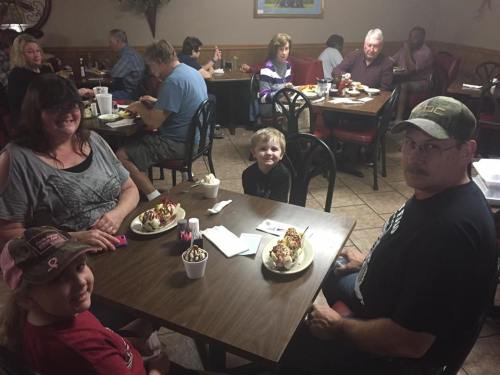 A family enjoys dessert at a restaurant, seated at a table with ice cream sundaes in front of them.