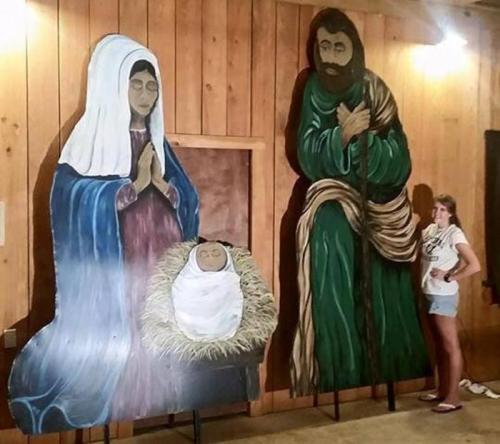 A girl stands next to large painted figures of Mary, Joseph, and baby Jesus in a rustic wooden setting.