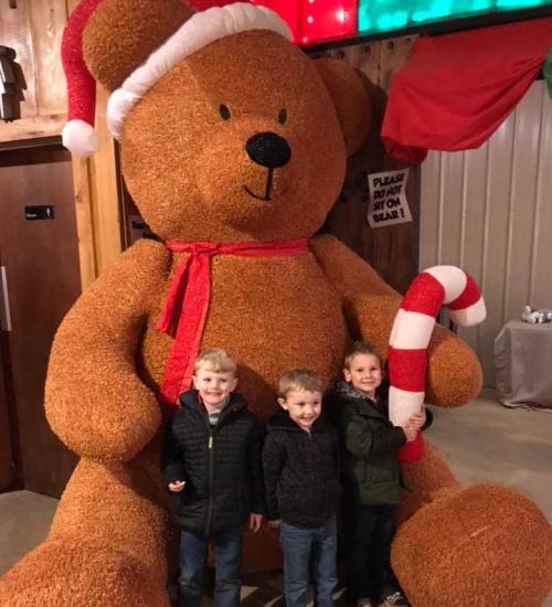 Three children stand in front of a large teddy bear wearing a Santa hat, holding a candy cane.