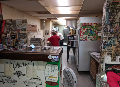 A busy pizzeria interior with a counter, kitchen area, and walls decorated with photos and posters.