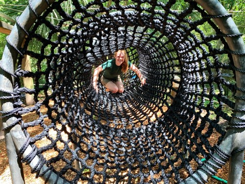 A person smiles while crawling through a large, circular rope net structure in a playground.