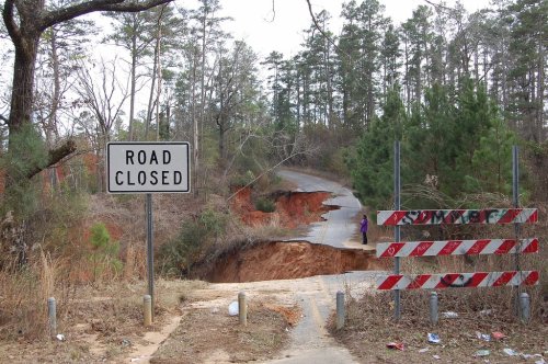 A "Road Closed" sign near a damaged road with a large sinkhole, surrounded by trees and a barrier.