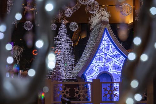A festive scene featuring a blue-lit house, decorated trees, and sparkling ornaments in a winter wonderland setting.