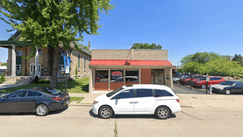 A small brick building with a red awning, parked cars in front, and trees in the background on a sunny day.