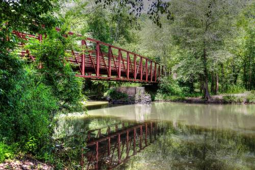 A red bridge spans a calm river, surrounded by lush green trees and reflecting in the water below.
