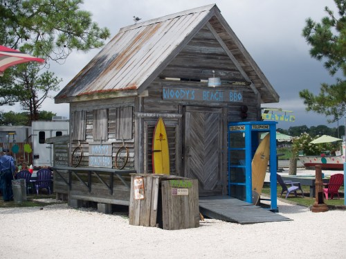 A rustic wooden building with a sign reading "Woody's Beach BBQ," featuring surfboards and a telephone booth outside.