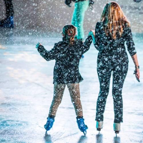 A woman and a child skate together on an ice rink, surrounded by falling snowflakes.