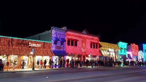 Colorful holiday lights adorn storefronts along a bustling street at night, with a crowd enjoying the festive atmosphere.