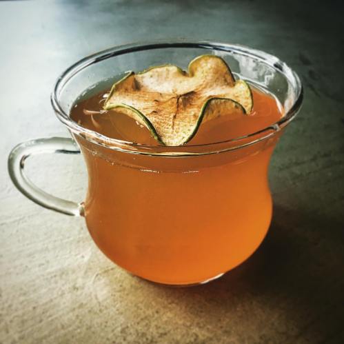 A clear glass cup filled with a warm, amber-colored drink topped with a dried apple slice.