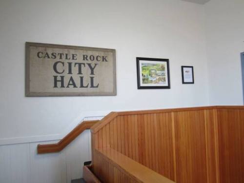 Interior view of a staircase leading to Castle Rock City Hall, featuring a sign and framed artwork on the wall.