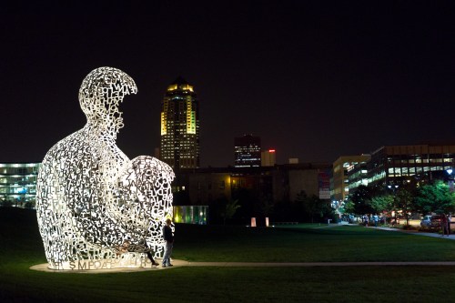 A large illuminated sculpture of a parent and child made of letters, set against a city skyline at night.