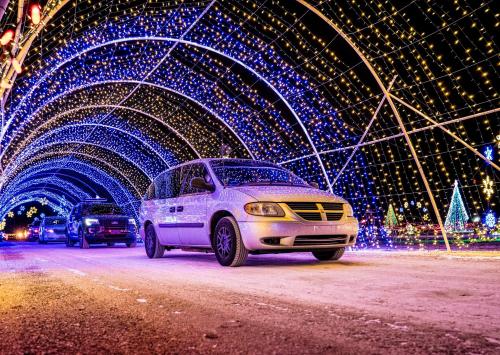 A white minivan drives through a colorful, illuminated tunnel of holiday lights with trees in the background.