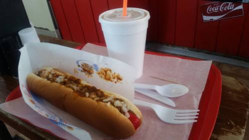 A hot dog topped with chili and onions on a tray, accompanied by a drink and utensils.