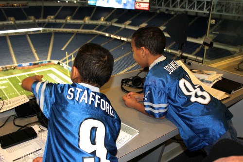 Two boys in blue football jerseys point out from a press box overlooking an empty stadium.