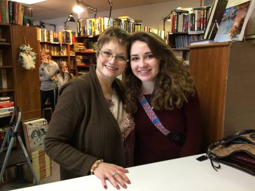 Two women smile together in a bookstore, surrounded by shelves of books. A few people are visible in the background.