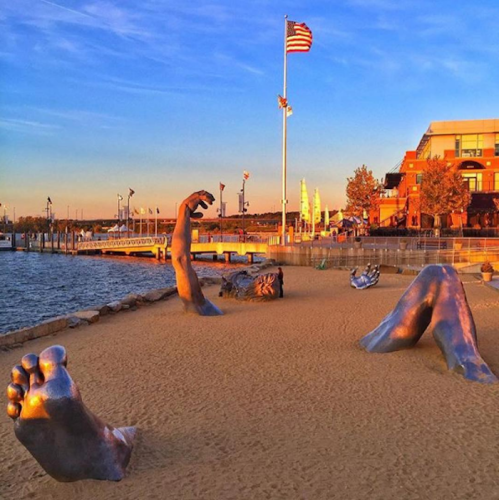 Sculptures of hands and feet on a sandy beach by the water, with a flag and buildings in the background at sunset.
