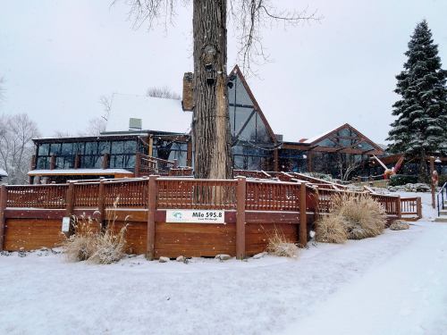 A large, modern lodge with a wooden deck, surrounded by snow and trees, on a winter day.