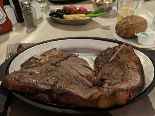 A large, grilled steak on a metal plate, accompanied by a baked potato and a side of olives and vegetables.