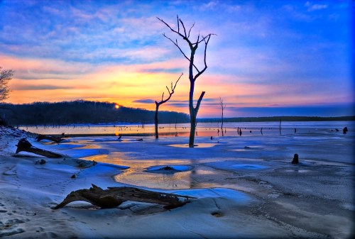 A serene winter landscape featuring a frozen lake, bare trees, and a colorful sunset reflecting on the ice.