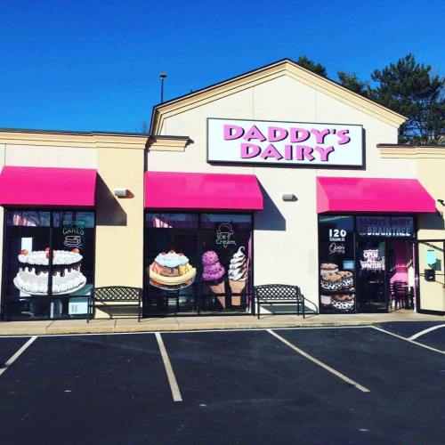 Exterior of Daddy's Dairy, featuring pink awnings and large images of ice cream and cakes on the windows. Clear blue sky.