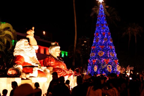 A brightly lit Christmas tree stands beside Santa and Mrs. Claus decorations, with a crowd enjoying the festive atmosphere.