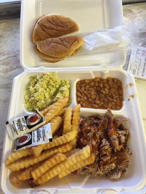 A takeout container with pulled pork, crinkle-cut fries, coleslaw, baked beans, and two sandwich buns with ketchup packets.
