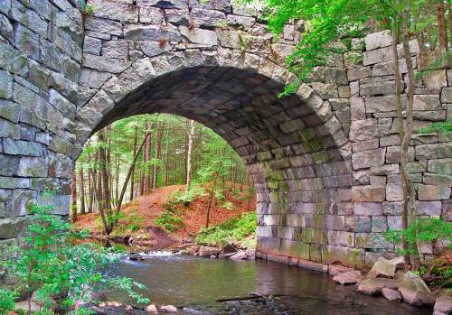A stone arch bridge over a calm stream, surrounded by lush green trees and a forested landscape.