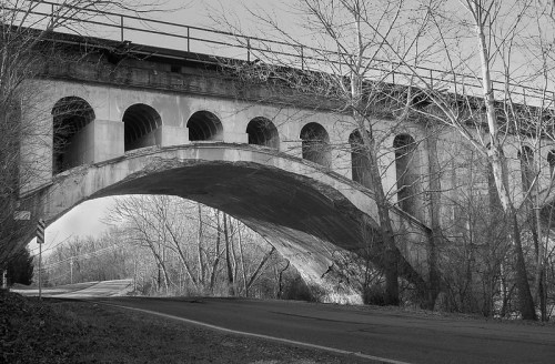 Black and white image of a large arch bridge with trees and a road beneath it.