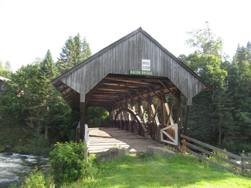 A wooden covered bridge named Bacon Bridge, surrounded by trees and a river, with a "Bridge Closed" sign.