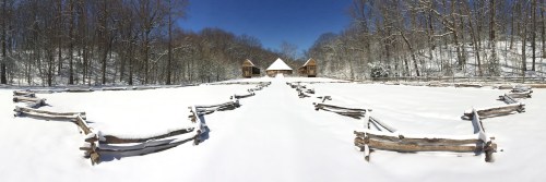 A snowy landscape with two wooden buildings and a fenced area, surrounded by trees under a clear blue sky.