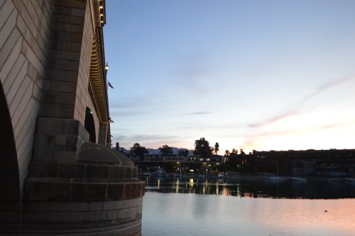 A serene sunset over a calm lake, with a bridge arching above and buildings reflecting in the water.